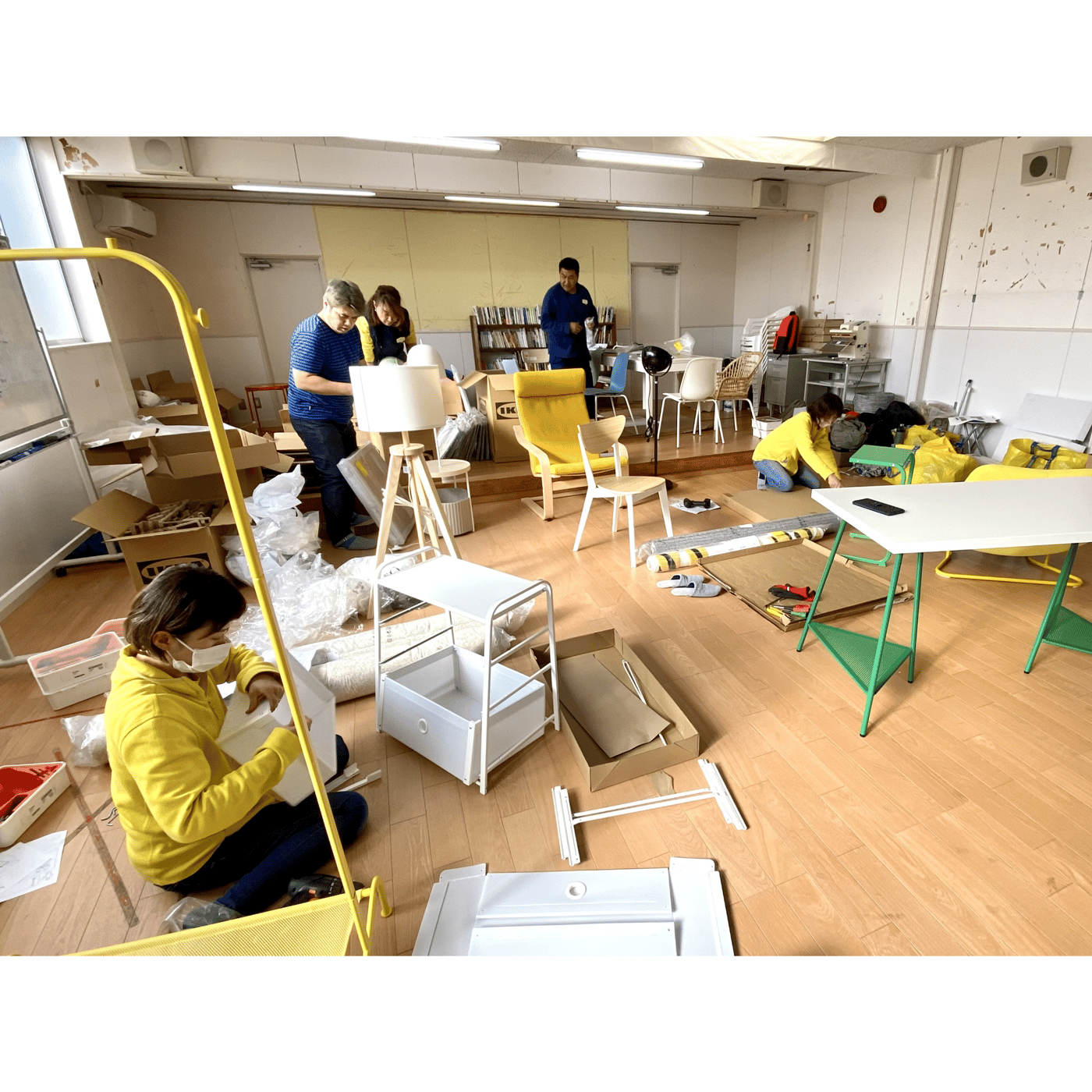黄色いシャツを着た人が、金属製の棚を組み立てている。奥にも数人家具を組み立てている。