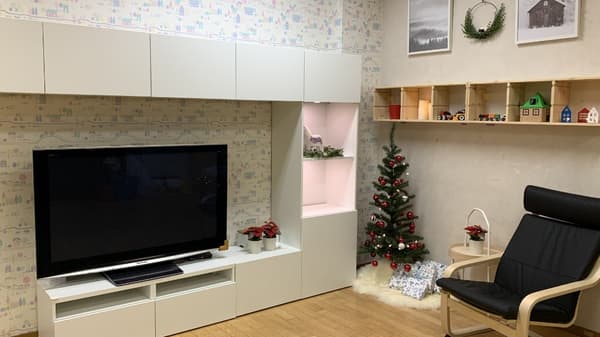 白いシェルフで囲われた壁際に、大型テレビが置かれている。部屋の角にクリスマスツリーが置かれ、黒い一人掛けソファーが置かれている。