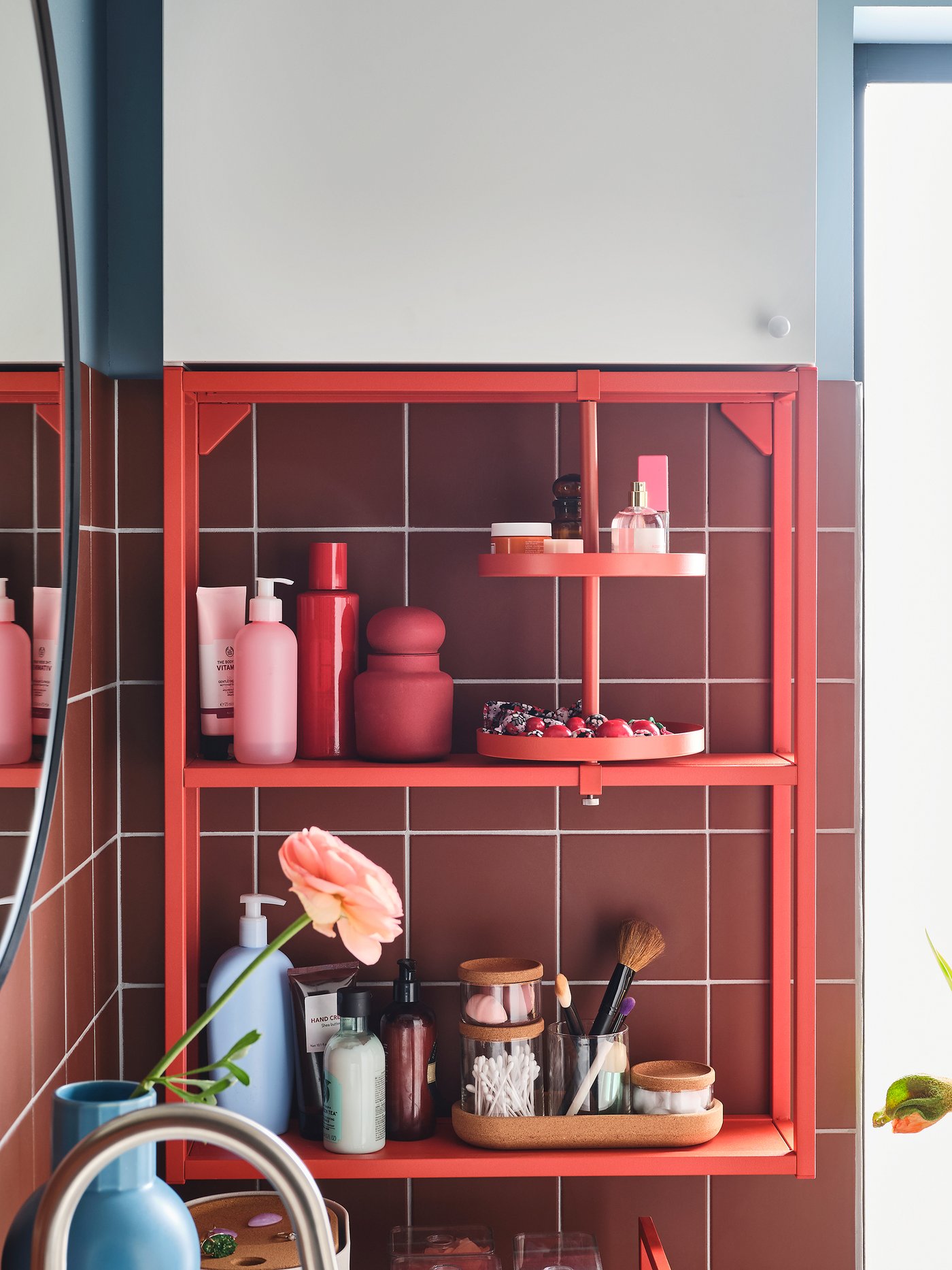 إطار جداري مع رفين ورف دوار باللون الأحمر البرتقالي، وخزانة حائط بيضاء، ومرآة مستديرة ومزهرية زرقاء.