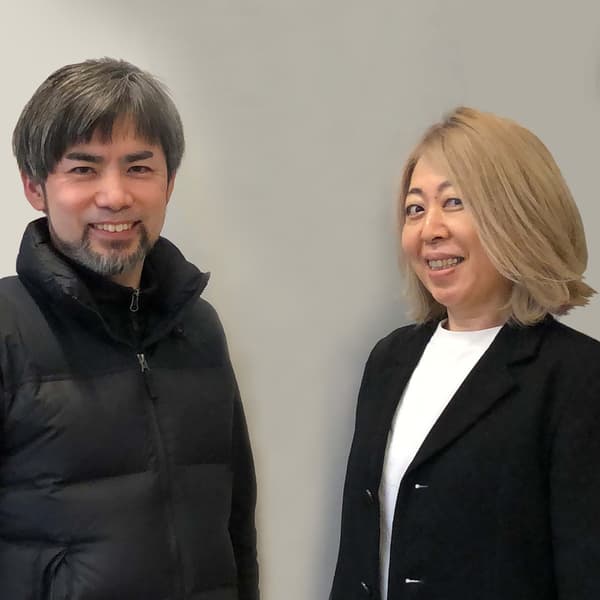 短い髪とひげの人物と、肩までの金髪の人物が並んで立っている。