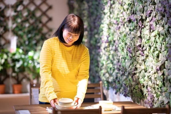 黄色いトップスの人物がテーブルウェアを並べ、グリーンウォールと鉢植えが映えるイケアのダイニング