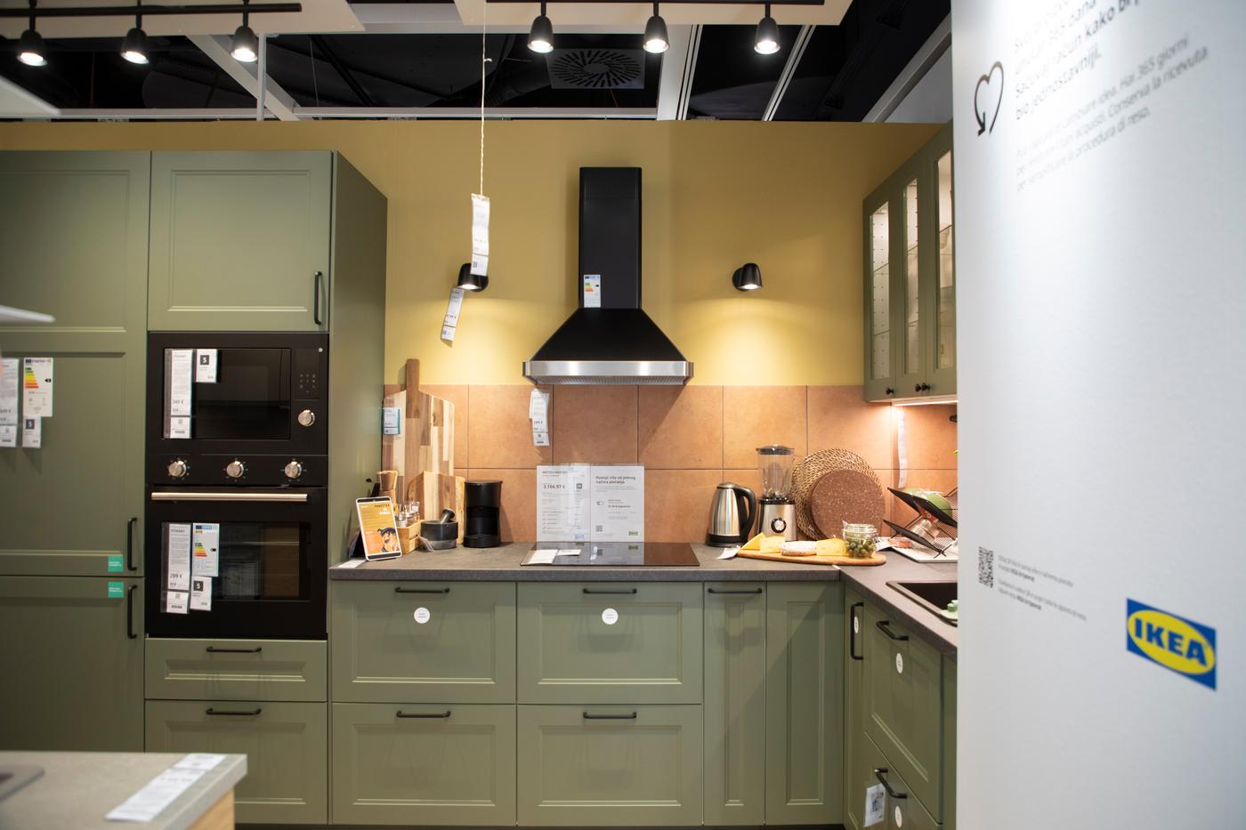 Izložbena kuhinja u IKEA studiju za planiranje s maslinastozelenim ormarićima, crnom napom i ugradbenom pećnicom, te kuhinjskim priborom i aparatima na radnoj površini.