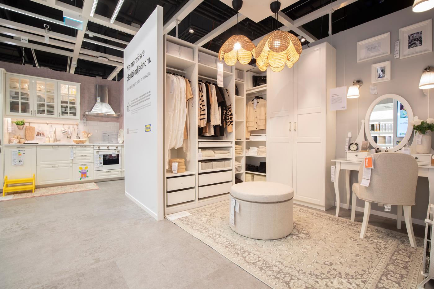 Izložbeni prostor IKEA studija za planiranje s bijelim ormarom PAX punim odjeće, toaletnim stolićem s ogledalom, tepihom i pufom, te kuhinjskim izložbenim kutkom u pozadini.