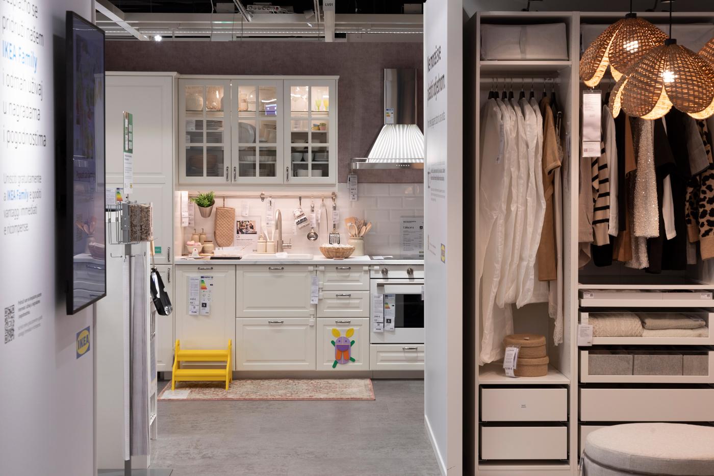 Izložbeni prostor IKEA studiju za planiranje s bijelom kuhinjom u pozadini i otvorenim ormarom PAX s obješenom odjećom i ladicama u prvom planu, te pletenom lampom.