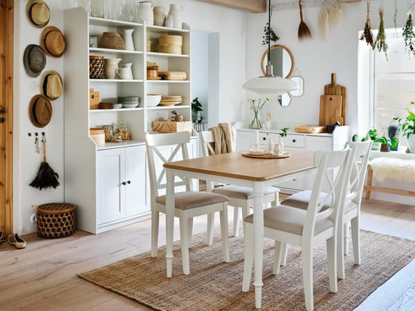 Jadalnia z białym stołem z dębowym blatem, czterema białymi krzesłami, jutowym dywanem i dwoma białymi, dwudrzwiowymi, wysokimi szafkami.