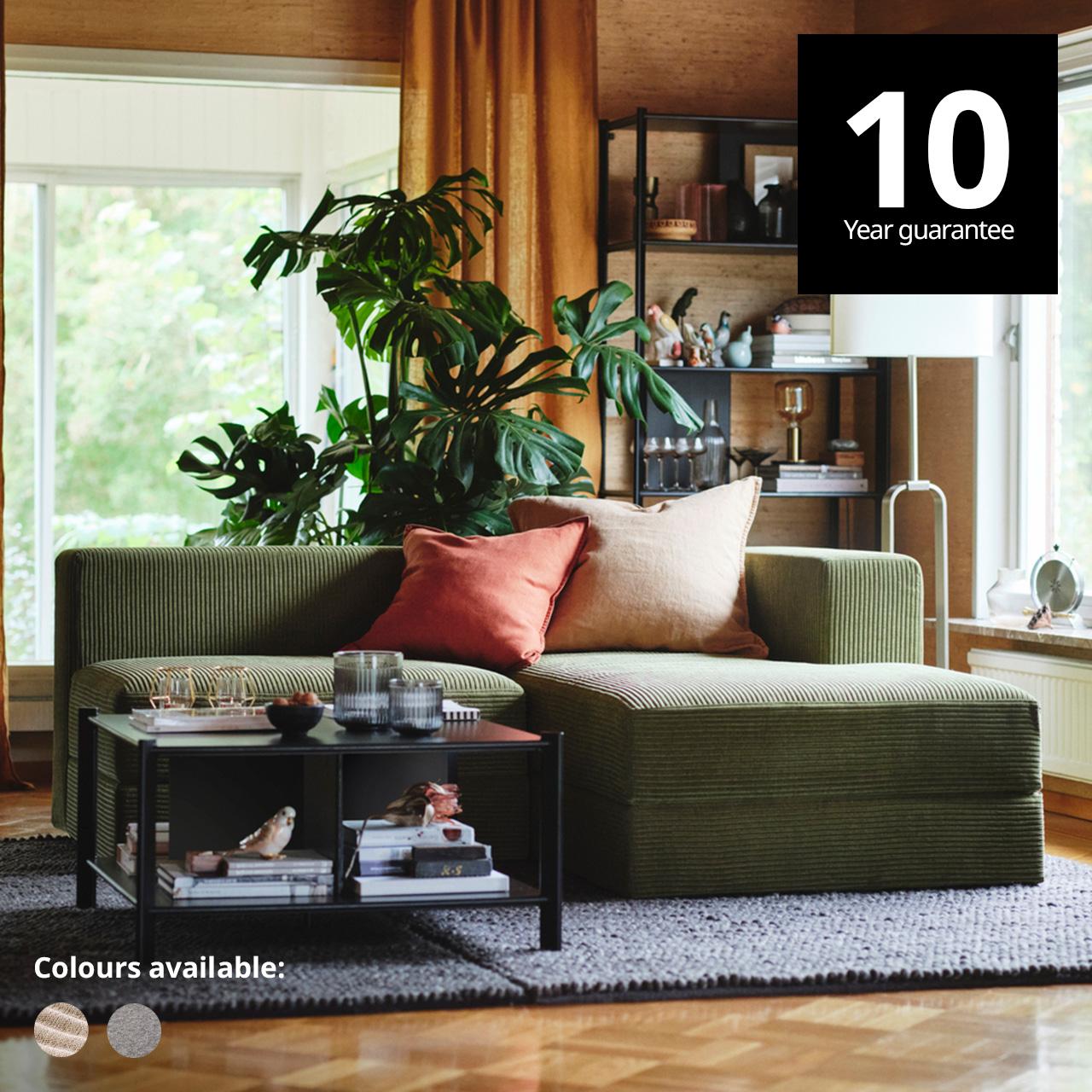 JÄTTEBO modular sofa in samsala dark yellow-green colour with GURLI salmon red and yellow cushion on top in spacious living room setting on top of a VONSBAK blue rug and a stockholm coffee table. The large windows behind and to the side of the sofa are framed with orange sheer curtains and a UPPVIND floor lamp stands in the corner with a shelf.  A 10-year guarantee tag is placed over the image at the top right corner. White text overlay image that reads "Colours available:" with the colour swatch of Samsal grey-beige and Tonerud grey.