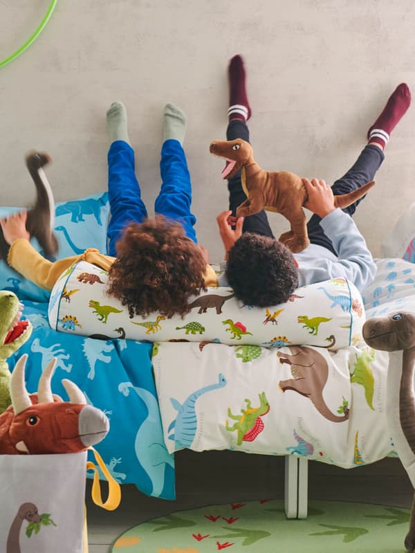 JÄTTELIK ágyneművel behúzott ágyon két gyerek fekszik, fejüket JÄTTELIK párnán nyu-gatják és dinoszauruszos JÄTTELIK puha játékokkal játszanak.