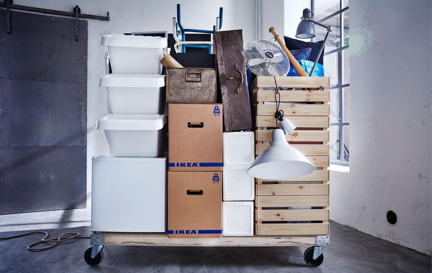 빈방에 다양한 이사용품과 JÄTTENE 예테네 상자가 쌓여 있는 바퀴 달린 목재 팔레트가 있습니다.