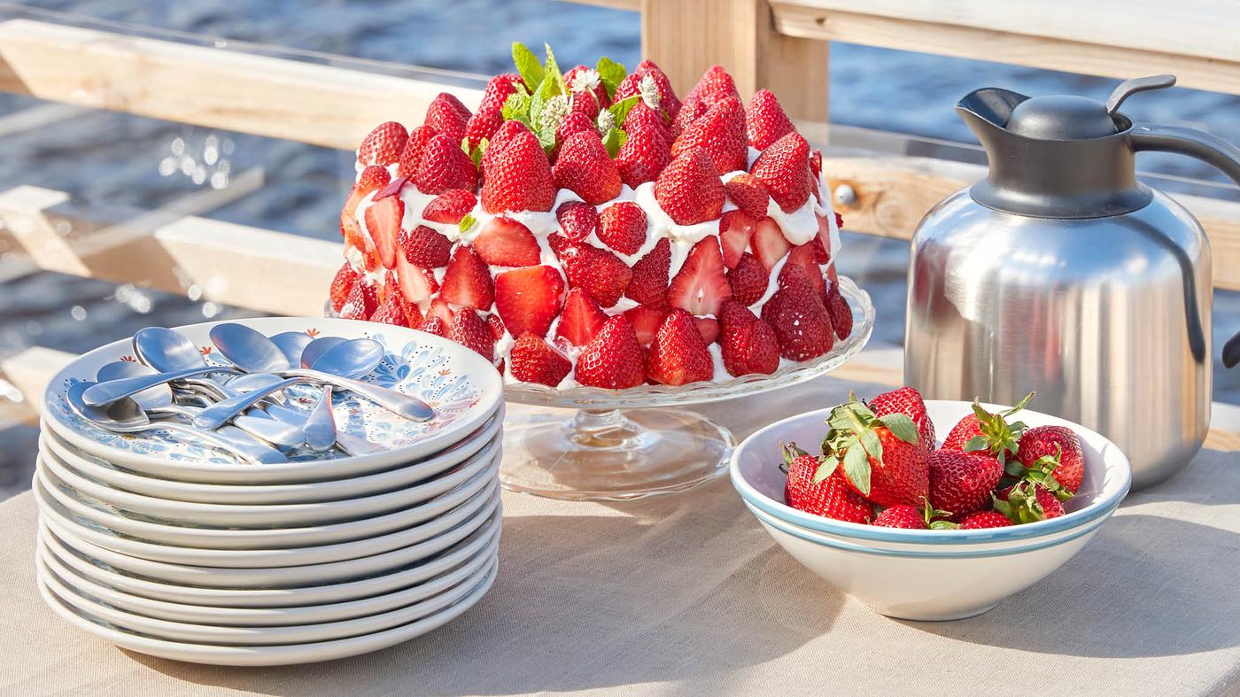 Jahodový dort na stojanu ARV BRÖLLOP na stole vedle dezertních talířů a misky jahod.