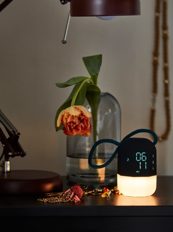 Jam loceng yang memancarkan cahaya hangat dan lembut dan vas kaca yang mengandungi bunga berwarna jingga terletak di atas meja sisi katil di dalam bilik tidur.