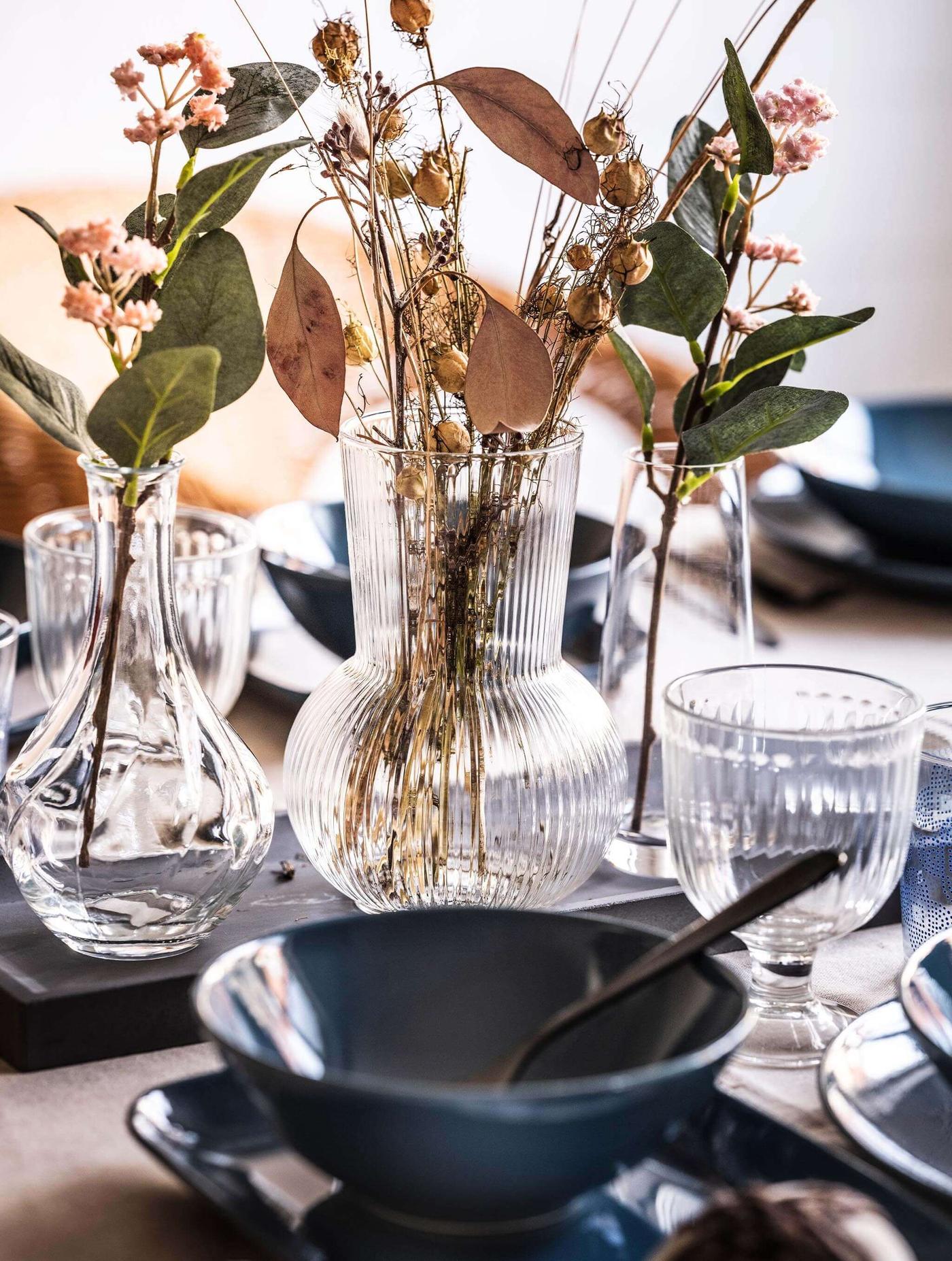 Jarrón de vidrio transparente con flores y hojas secas sobre una mesa. Delicados tallos y pétalos en tonos café y pasteles apagados.