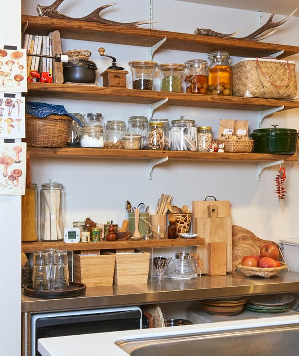 Jars of ingredients and pickles on wooden shelves above a metal worktop with wooden boxes and chopping boards.