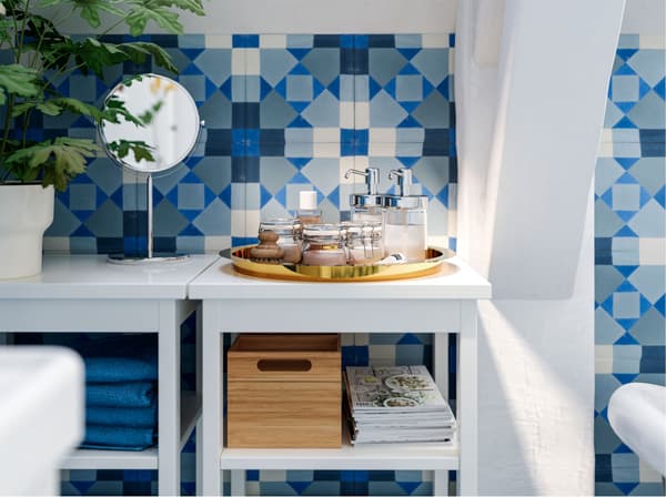 Jars, soap dispensers, a plant and a mirror on top of open white shelving units in a bathroom tiled with graphic coloured tiles.