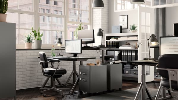 Jasne biuro ze stanowiskami pracy, które stoją naprzeciwko siebie. Fotel biurowy ALEFJÄLL stoi obok biurek IDÅSEN z regulacją wysokości oraz komód.