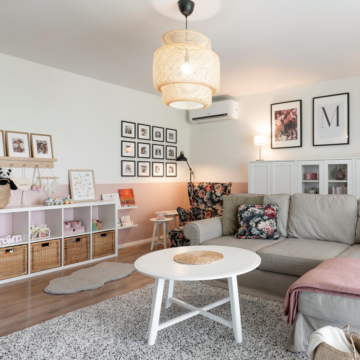 Jasne pomieszczenie z plecioną lampą sufitową, białym, okrągłym stolikiem, szarą kanapą i licznymi obrazkami na ścianach.