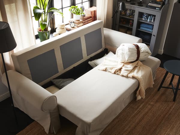 Jasnobeżowa sofa w trakcie składania po spaniu. W pojemniku pod siedziskiem leżą poduszki.
