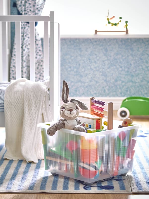 Játékokkal teli átlátszó VESSLA dobozt látunk egy fehér és kék csíkos szőnyegen. A hát-térben egy SUNDVIK rácsos ágy áll.