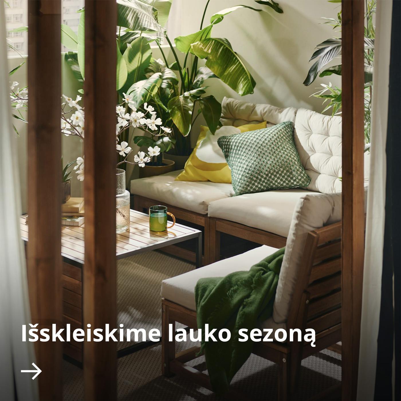 Jaukus, saulės nušviestas poilsio kampelis balkone su vazoniniais augalais, mediniais baldais, smėlinės spalvos pagalvėlėmis ir žaliais akcentais.