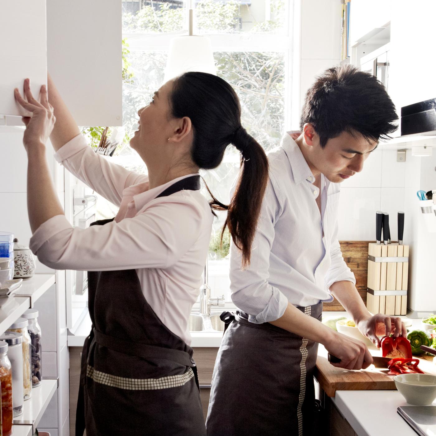 Jeune couple préparant à manger dans une cuisine linéaire. L’un d’eux hache un poivron tandis que l’autre a le nez dans une armoire murale.