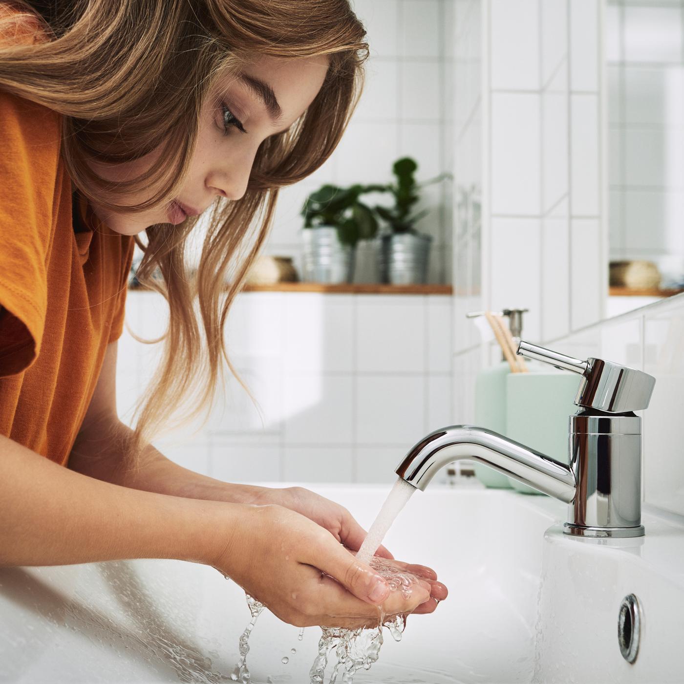 Jeune enfant aux longs cheveux brun se lavant les mains dans le lavabo d’une salle de bain carrelée de blanc 