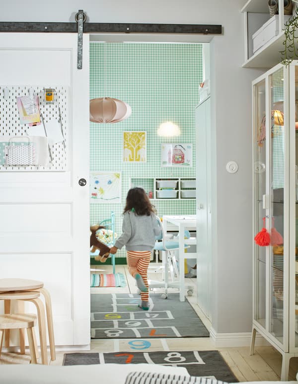 Jeune enfant passant en courant la porte coulissante ouverte d’une pièce blanche et verte meublée d’un lit, d’un espace de rangement, d’un bureau et plus encore.