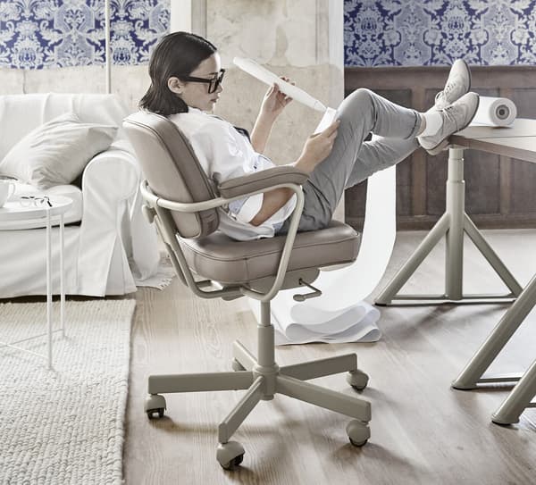 Jeune femme, assise sur une chaise beige, les pieds sur le bureau, dans un espace décoré de papier peint à motif bleu.