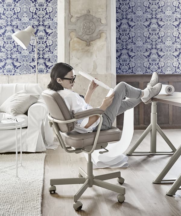 Jeune fille appuyée en arrière sur une chaise pivotante beige, avec les pieds sur le bureau, dans une pièce décorée d'un papier peint bleu à motifs.