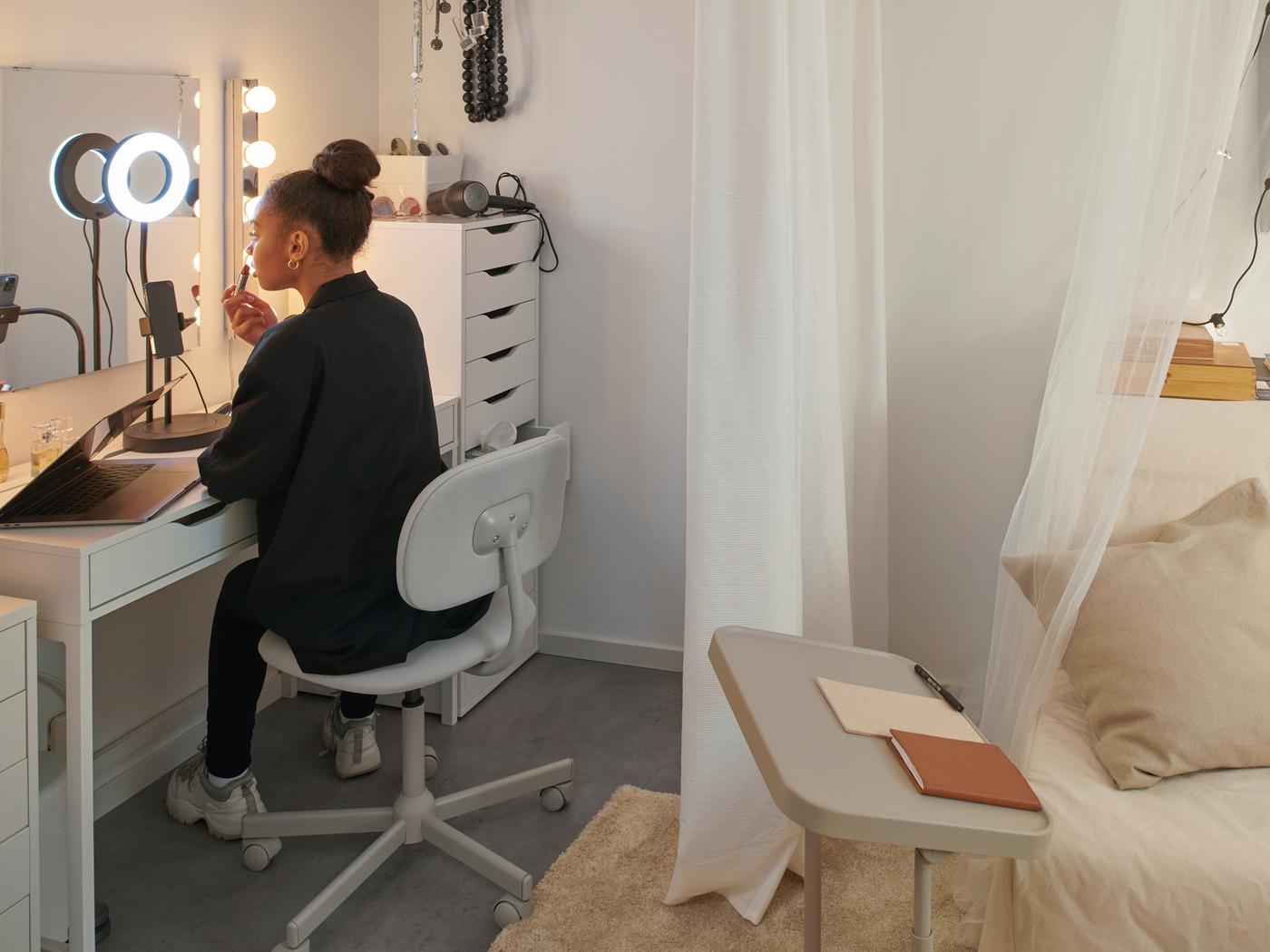 Jeune fille dans sa chambre d’étudiant assise sur une chaise pivotante BLECKBERGET et appuyée sur un bureau MICKE. Elle se maquille et un anneau lumineux LÅNESPELARE est allumé.