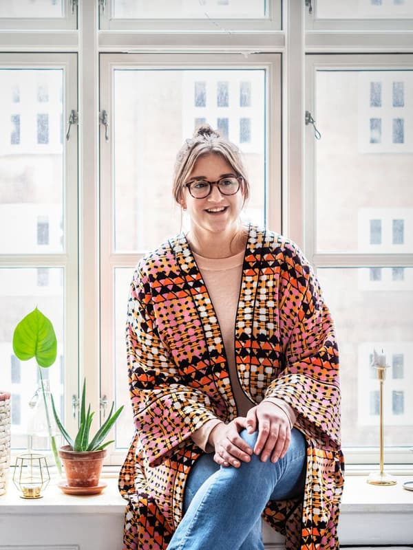 Jeune fille souriante vêtue d’un caftan très coloré, assise sur un appui de fenêtre de son appartement.