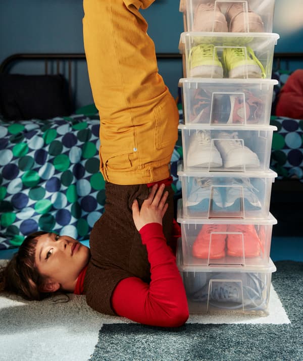 Jeune fille tenant la position de la chandelle à proximité d’une pile de boîtes SAMLA contenant des chaussures colorées.