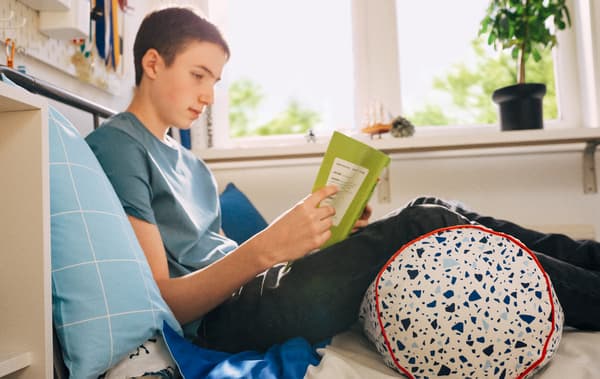 Jeune garçon en train de lire, assis sur son lit, le dos appuyé contre un oreiller et les jambes étendues au-dessus d’un coussin MÖJLIGHET.