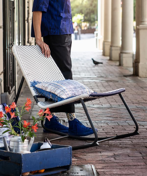 Jeune homme debout à côté d’une chaise berçante ÖVERALLT bleu et blanc à structure en métal noir.