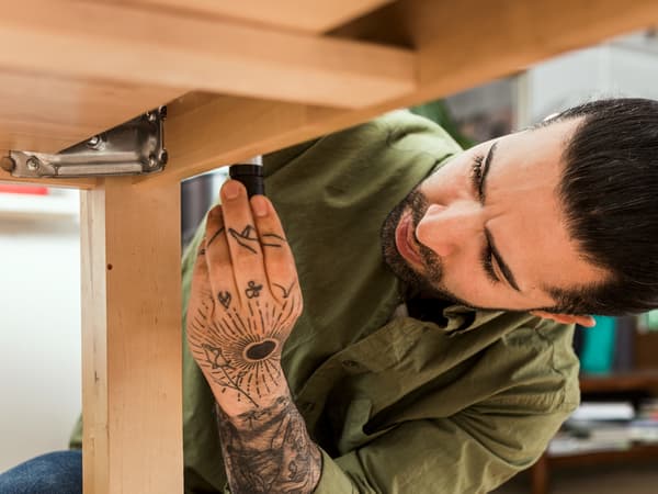 Jeune homme montant une table. En position accroupie, il est vêtu d’une chemise verte et d’un jean bleu. Son bras gauche est fortement tatoué.
