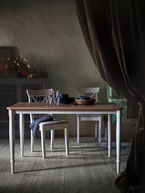 Jídelní stůl DANDERYD s dubovou dýhovou deskou a bílými nohami umístěný v temné místnosti s bílými židlemi.