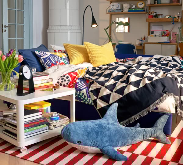 JOHANNE takaró, dekoratív mintával egy kanapé felett, lámpával, éjjeliszekrénnyel és sok könyvvel.