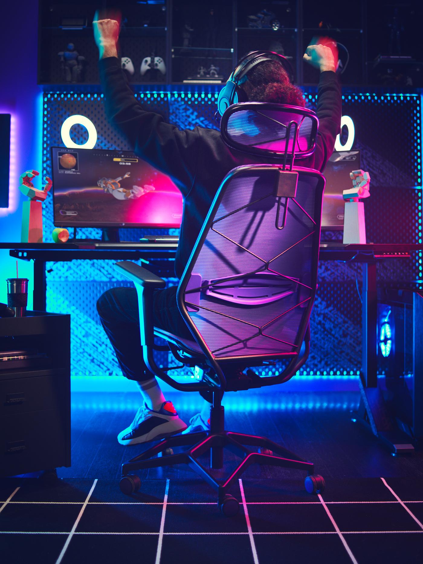 Jokatzeko gela iluna gaming-espazio argiztatu batekin. Pertsona bat, gaming-aulki batean eserita, besoak altxatzen, garaipenaren seinale.