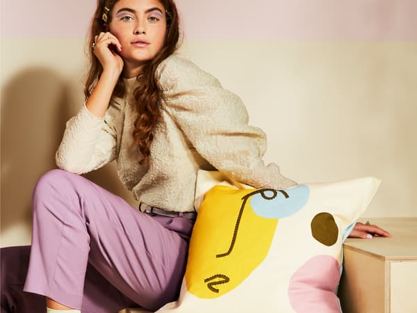 Jovem sentada numas escadas de madeira com uma almofada com um padrão abstrato de um rosto com cores vivas.