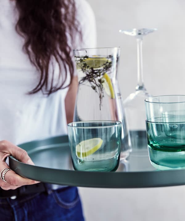 Julia tenant un plateau vert foncé avec des verres verts et une carafe d'eau avec du citron et des herbes.