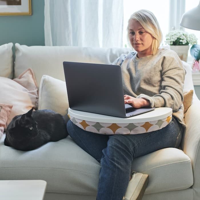 Junge Frau sitzt auf dem Sofa und hat einen Laptop mit Laptophalter auf den Knien.