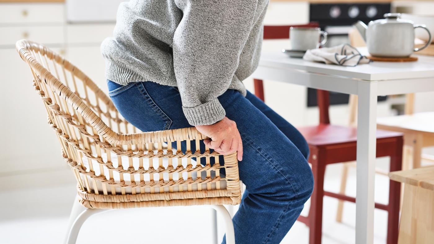 Junto a una mesa MELLTORP blanca, una persona con vaqueros y un jersey gris se levanta de una silla NILSOVE/NORNA de ratán con un cojín de silla.