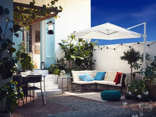 JUTHOLMEN sofa on a patio under a large umbrella with blue and red pillows. Plants on an outdoor setting.