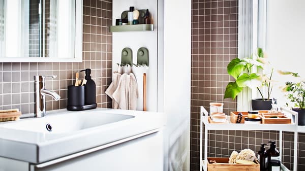 Kabinet bawah sink berwarna putih, cermin berwarna putih, troli berwarna putih dengan dulang dan balang selain kabinet tinggi berwarna putih di dalam bilik mandi berjubin berwarna kelabu.