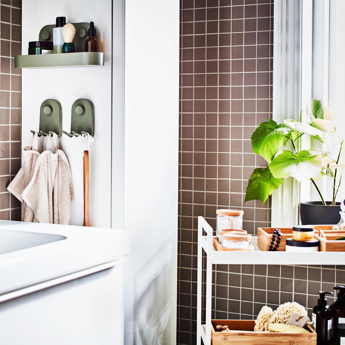 Kabinet bawah sink berwarna putih, troli berwarna putih dengan dulang dan balang, kabinet tinggi berwarna putih dan tumbuhan berpasu di dalam bilik mandi berjubin berwarna kelabu.