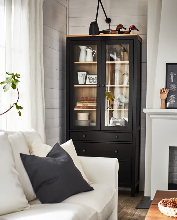 Kabinet berpintu kaca berwarna hitam coklat/kayu pain padu dengan item-item hiasan, lampu dinding berwarna hitam di atasnya dan sofa berwarna putih di hadapannya.