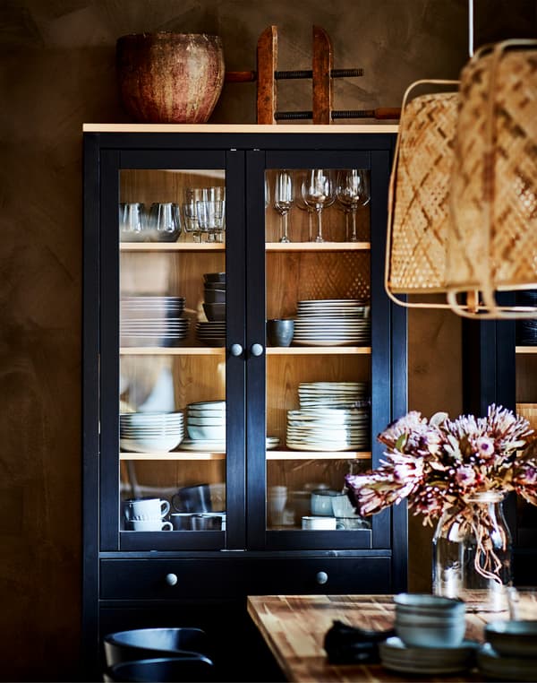 Kabinet berpintu kaca HEMNES berwarna hitam coklat yang mempamerkan gelas wain dan set makan malam, terletak di belakang meja makan yang mempunyai vas bunga.