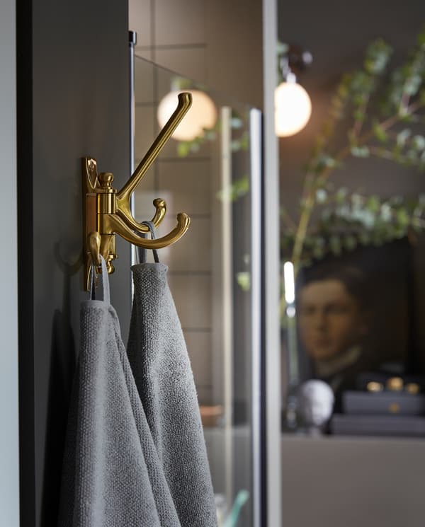 KÄMPIG 3-armed swivel hook in brass-colour is mounted on a wall next to a shower, and grey towels hang here to dry.