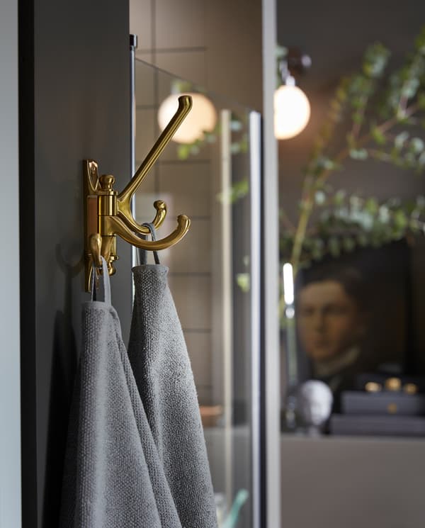 KÄMPIG 3-armed swivel hook in brass-colour is mounted on a wall next to a shower, and grey towels hang here to dry.