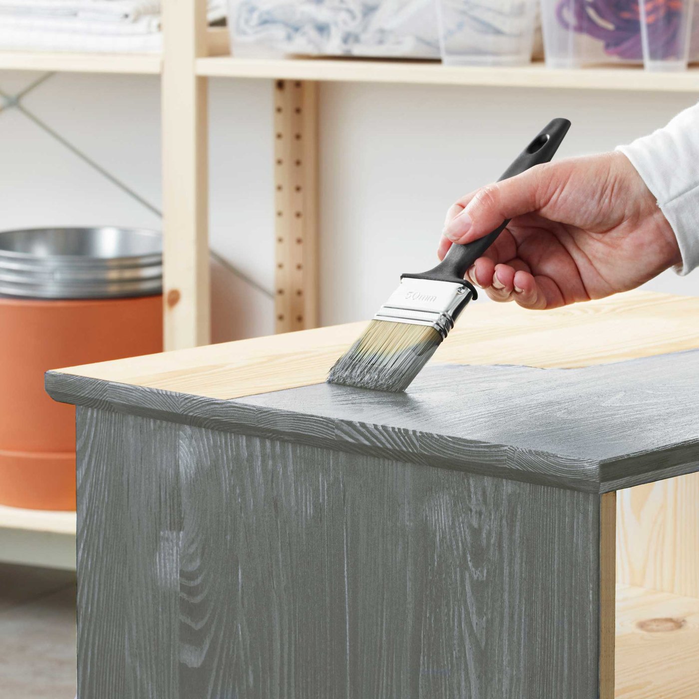 Käsi pitelee maalisivellintä ja maalaa käsittelemätöntä mäntykalustetta harmaalla maalilla.