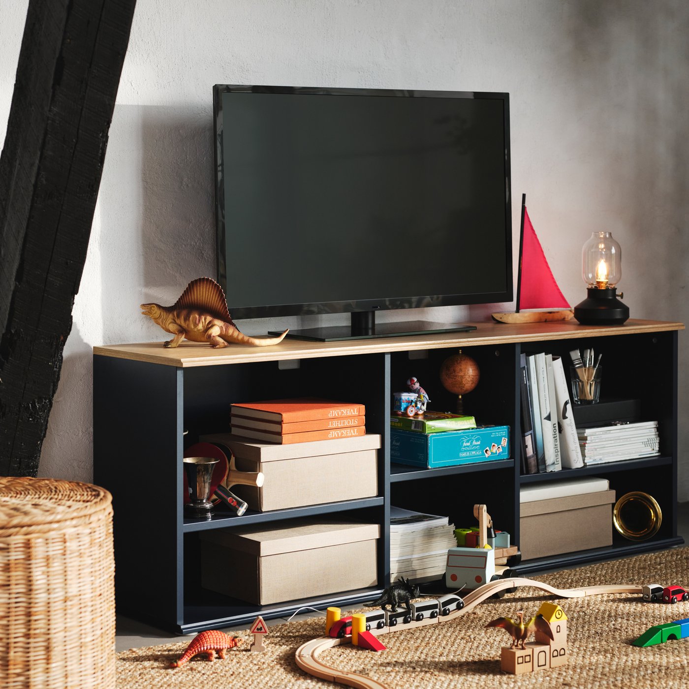 Kästen, Bücher und Spielzeug sind in den offenen Regalen einer schwarzblauen SKRUVBY TV-Bank zu sehen, auf der sich ein schwarzer Fernseher befindet.