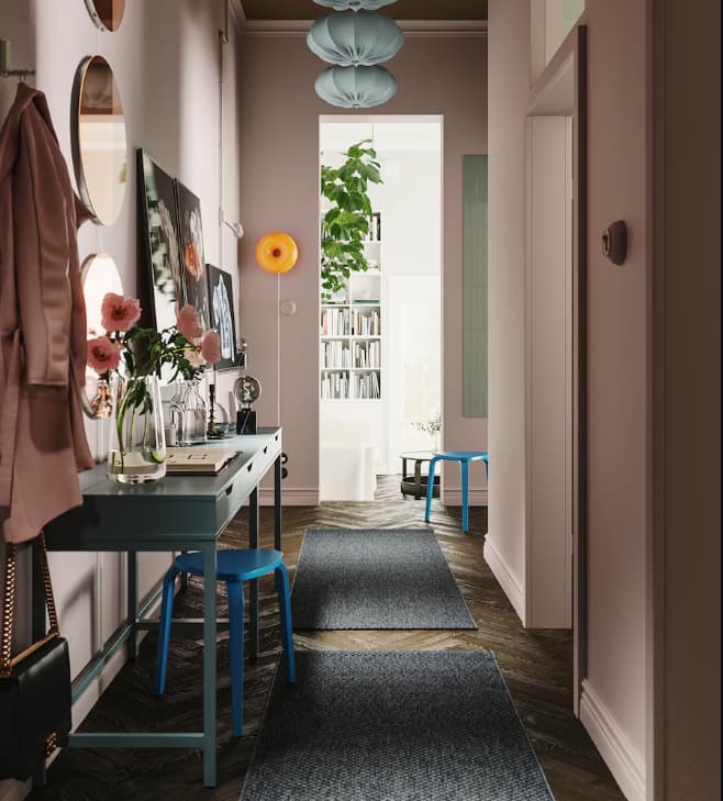 Käytävän seinää vasten on asetettu kaksi harmaanturkoosia ALEX-työpöytää. Toisen pöydän alla on KYRRE-jakkara. Katossa riippuu neljä REGNSKUR-varjostinta.